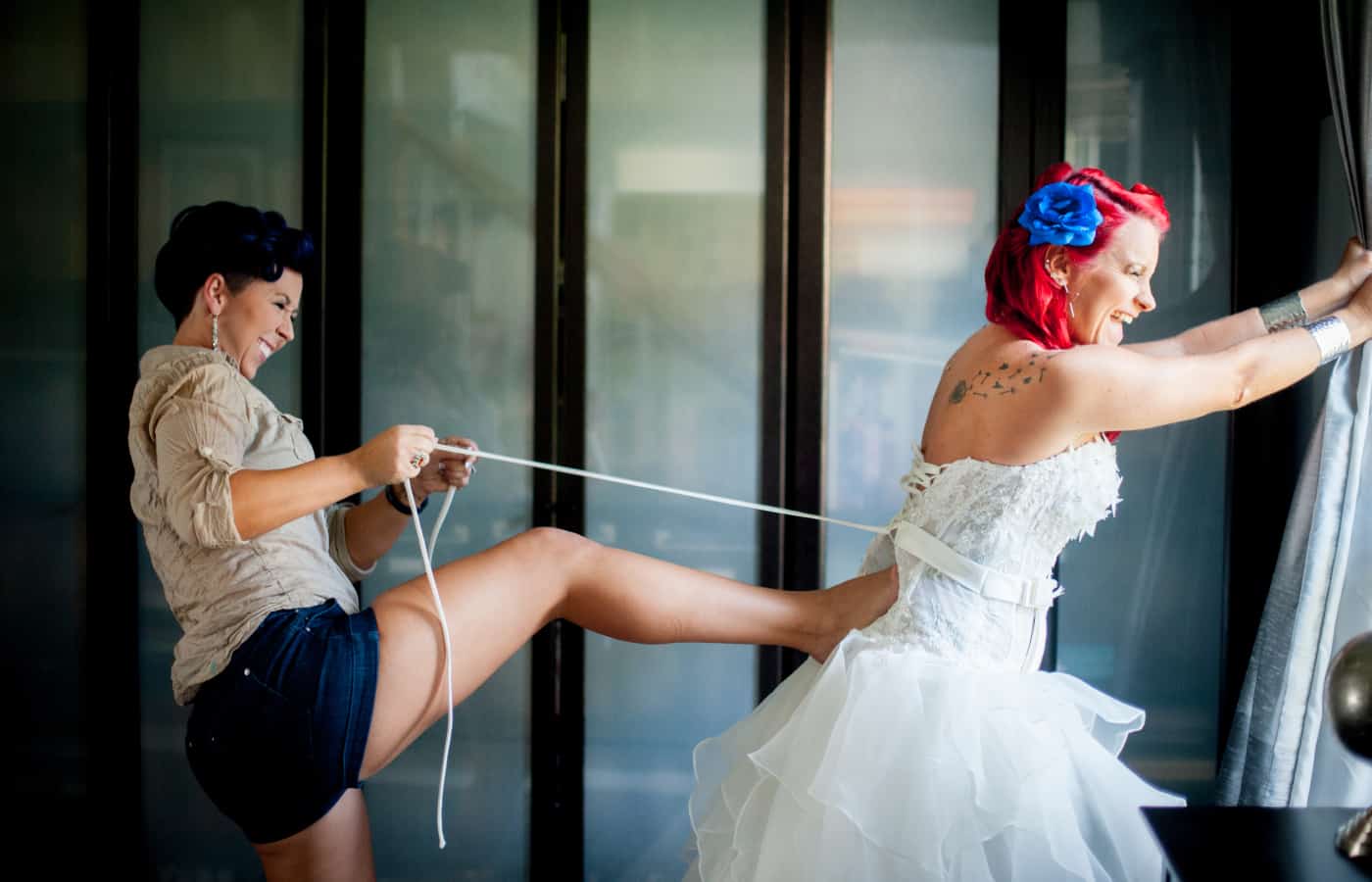 A bridemaid ties the back of a pink-haired bride's dress