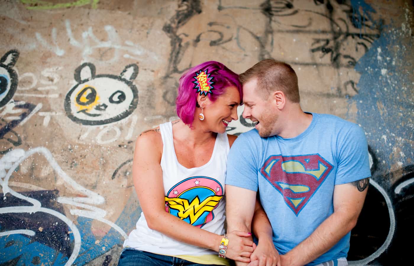 Graffiti background behind a woman with pink hair and a wonder woman t-shirt smiling at a man with a superman t-shirt
