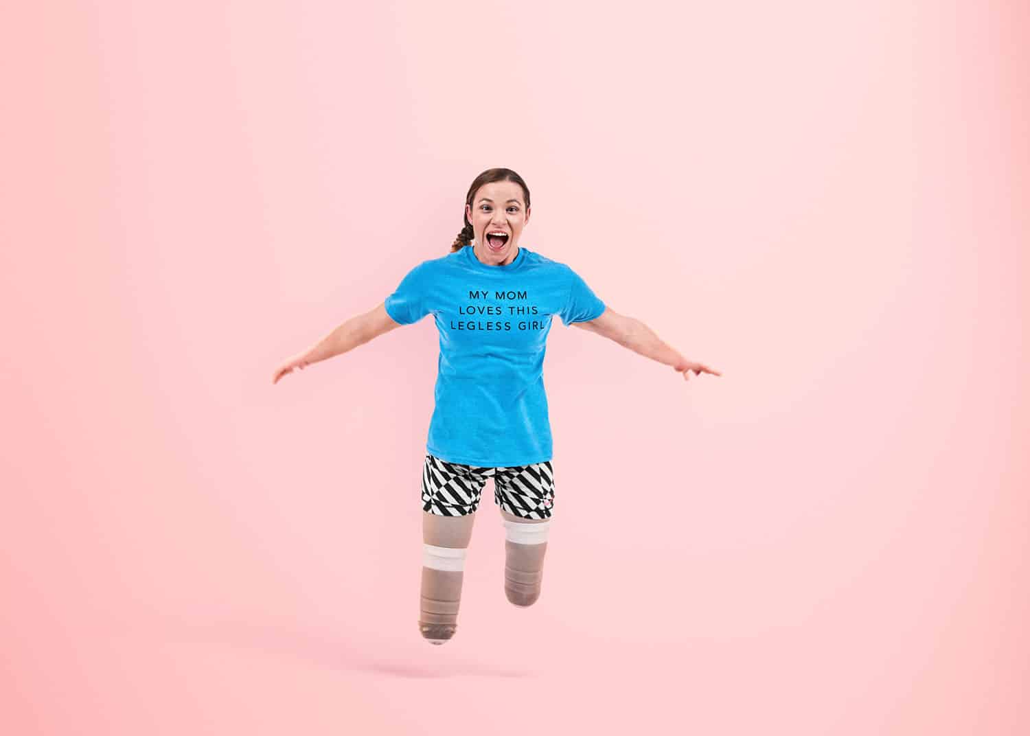 Paralympic gold medalist Oksana Masters leaps without her prosthetics on a pink background wearing a tee-shirt that says "My Mom Loves This Legless Girl."