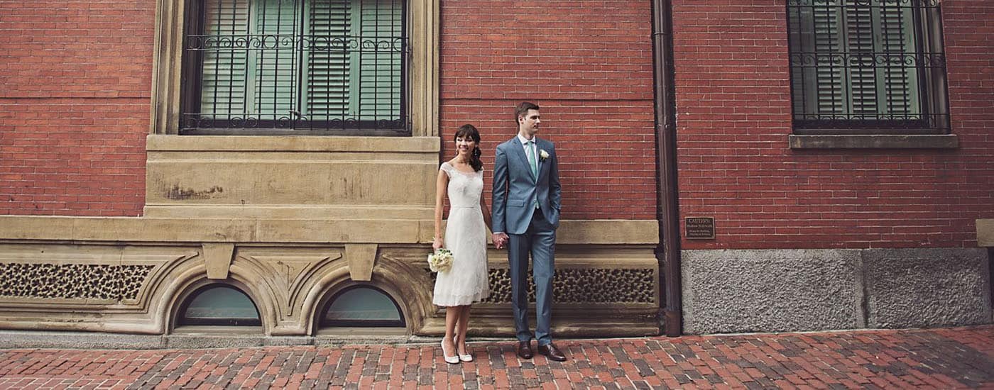 Bride and groom clasp hands in front of a tall brick wall. By Harris & Co.