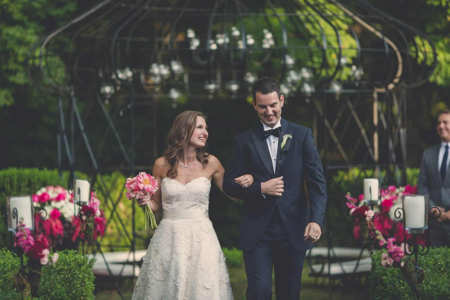 Bride and groom process out of their garden ceremony surrounded by pink flowers and greenery. By Harris & Co.