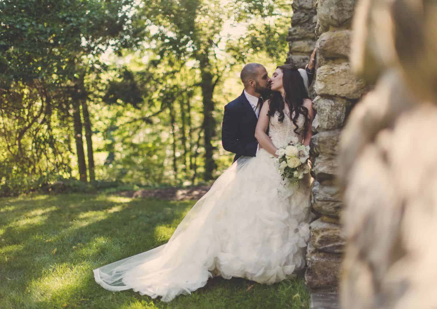 Groom kisses bride in ballgown against stone wall. By Harris & Co.