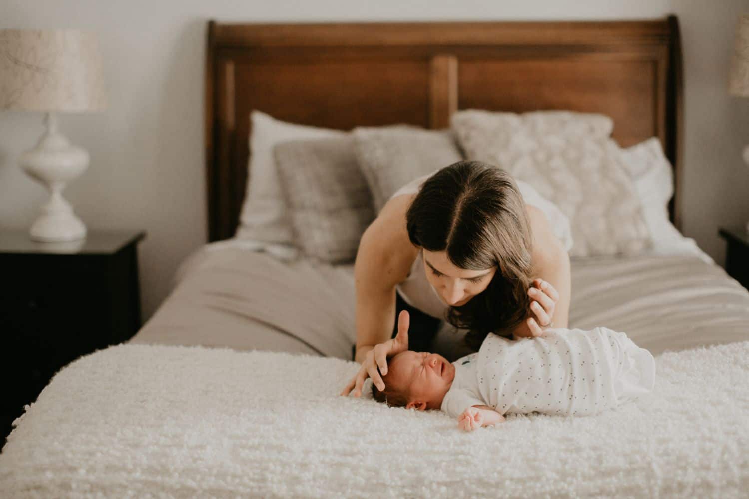 Lifestyle Newborn Photos: A swaddled newborn baby lies at the foot of their parents' bed as Mom kneels over the infant whispering words of comfort.