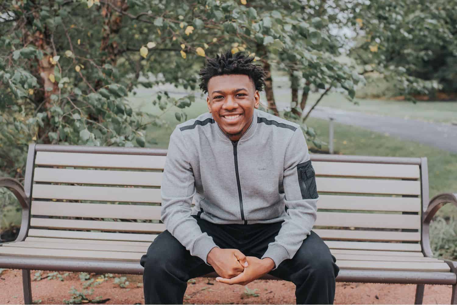 A handsome Black teen sits on a park bench smiling at the camera