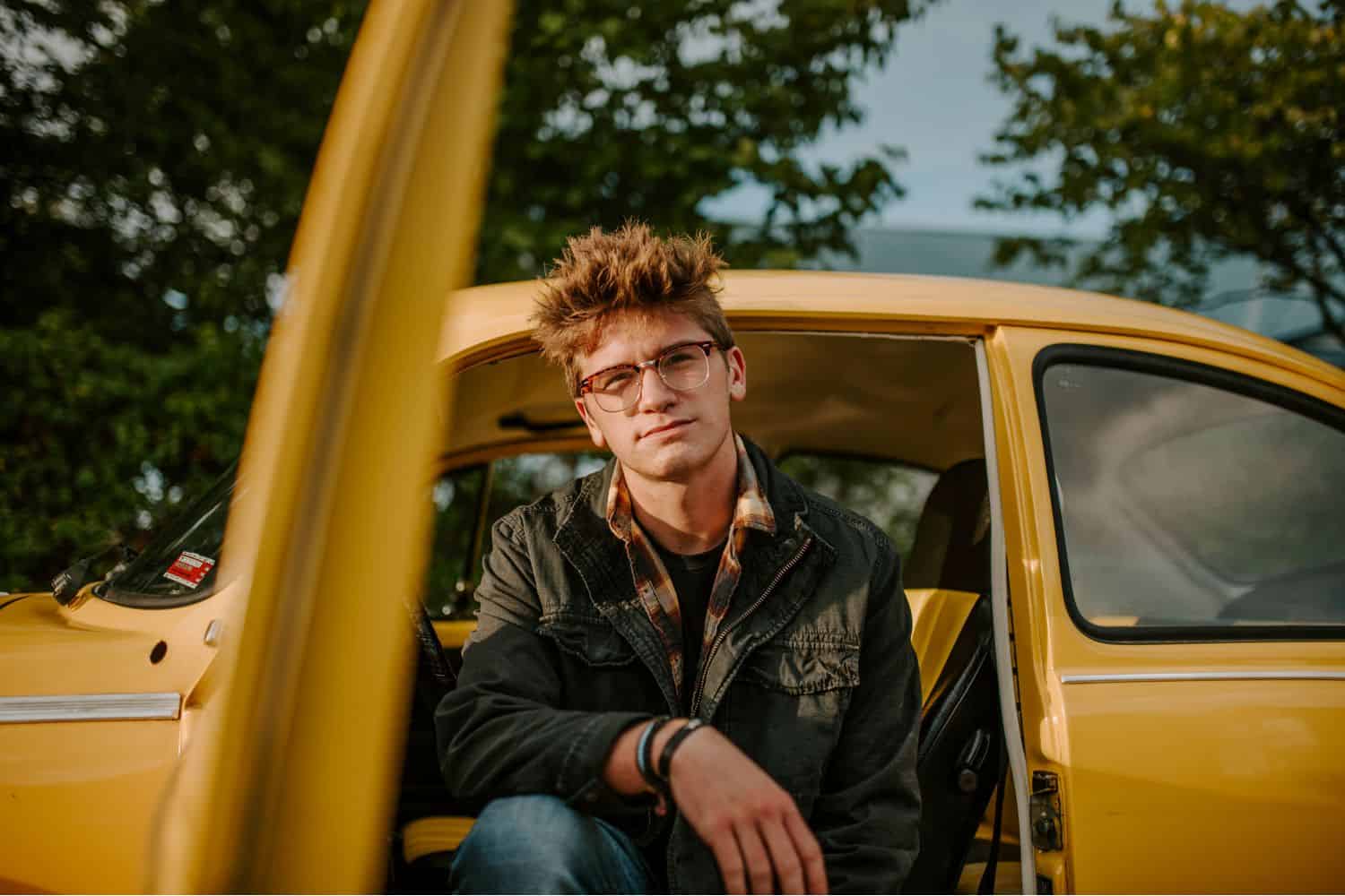 A high school senior guy sits in the open doorway of a bright yellow vintage car 