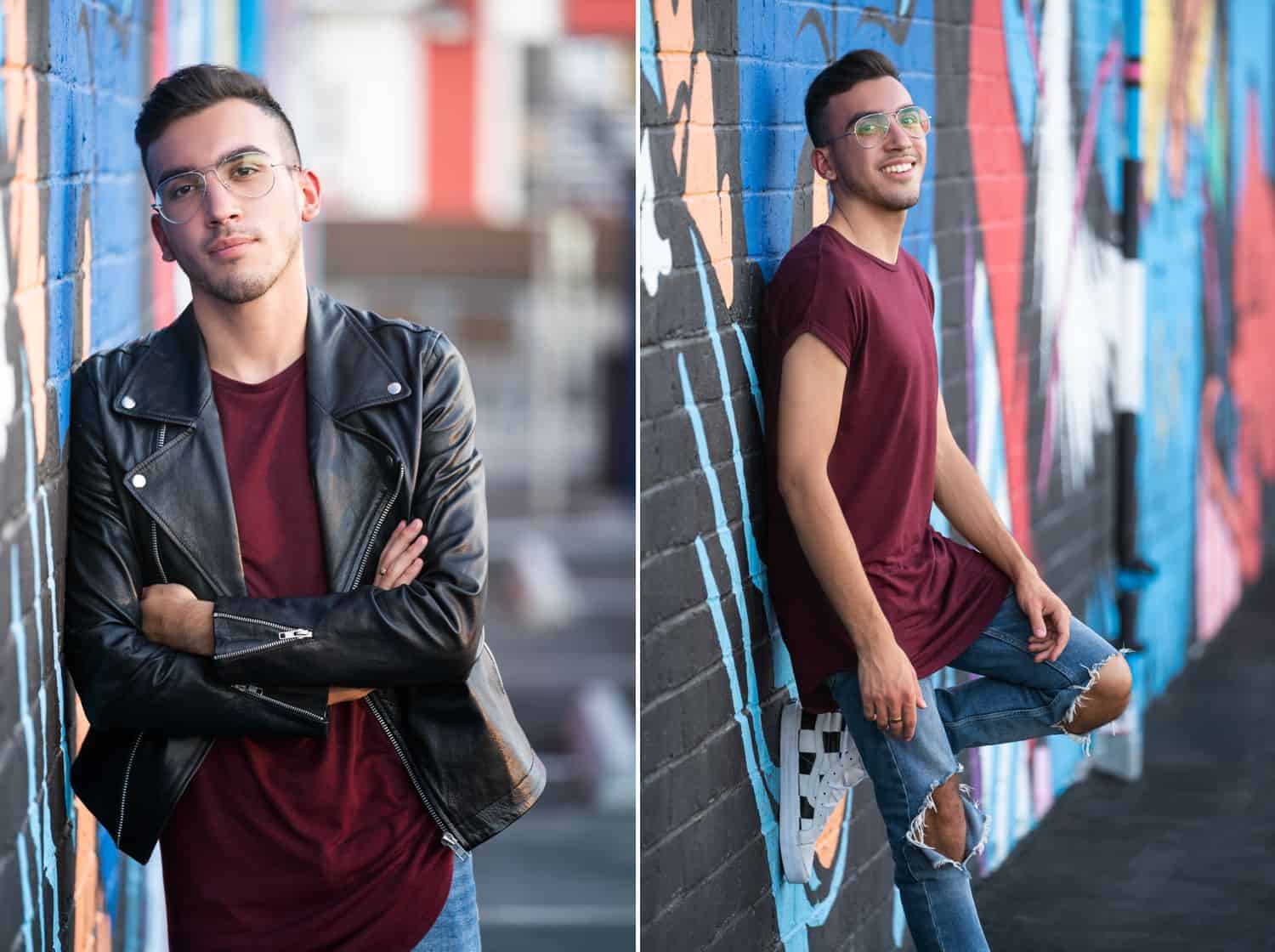 A guy wearing clear frame glasses and a leather jacket poses against a graffiti wall for his senior pictures.