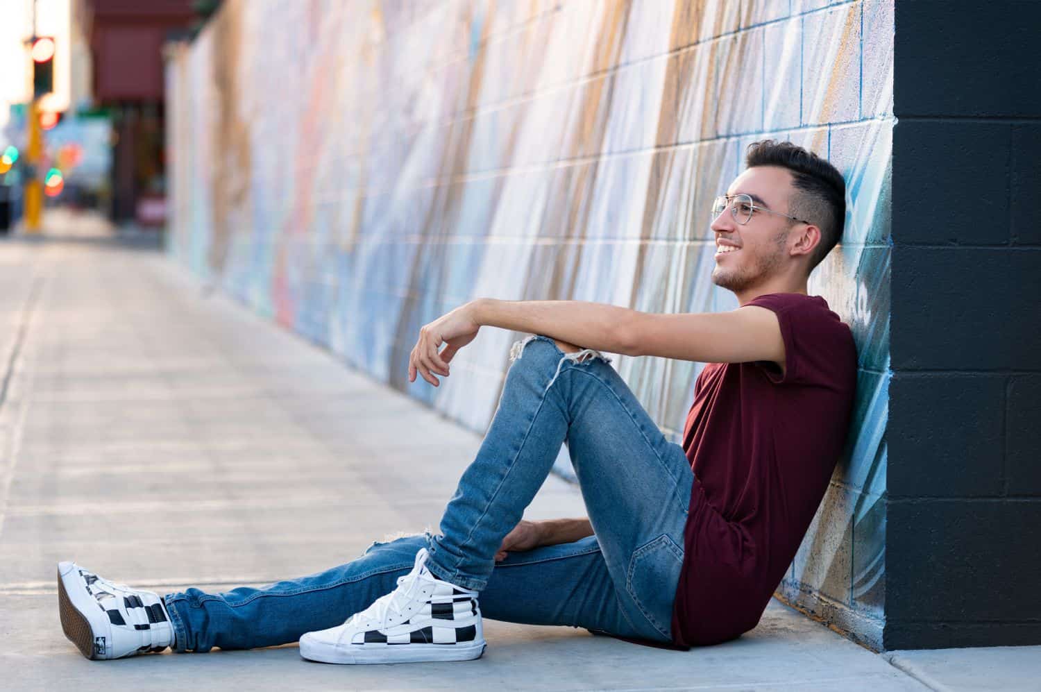 A high school senior boy wearing skinny jeans and checkerboard shoes leans against a graffiti wall and smiles into the distance.