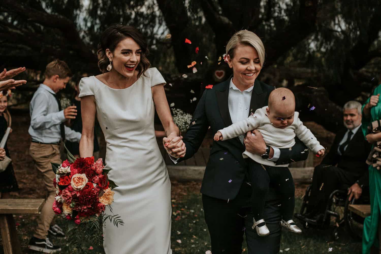 Two brides walk up the aisle with their baby amid a shower of rose petals