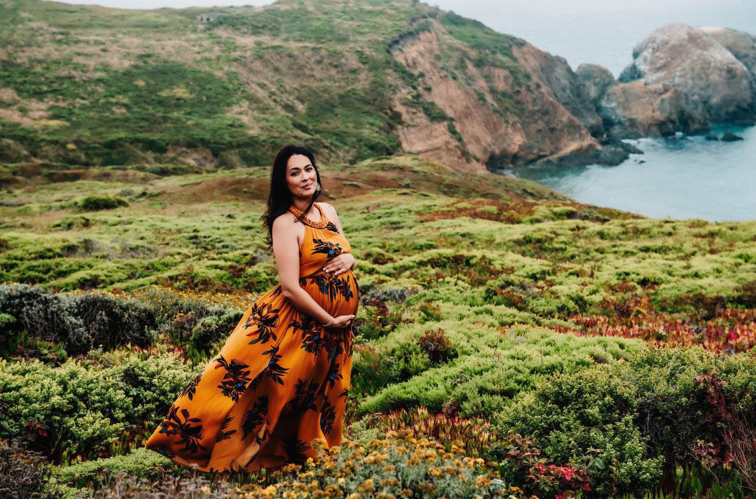 Captured Happiness Photography uses maternity poses like this one, which a pregnant woman standing on the seaside cliffs in Hawaii as her colorful dress flows behind her