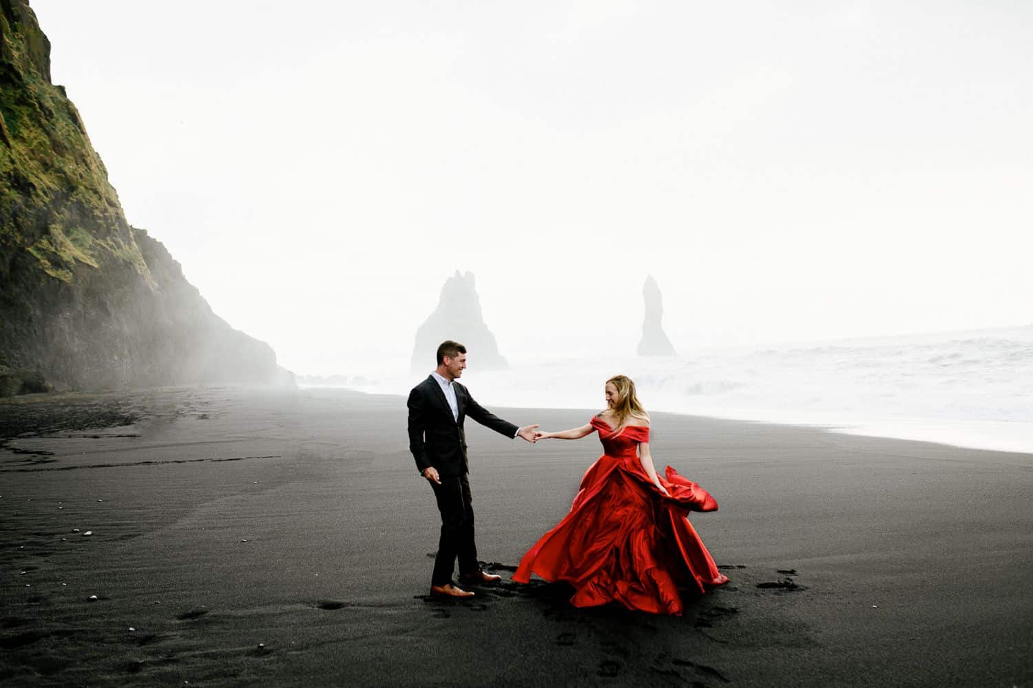 Man in a suit dances with woman in a red dress on the black sands of Iceland