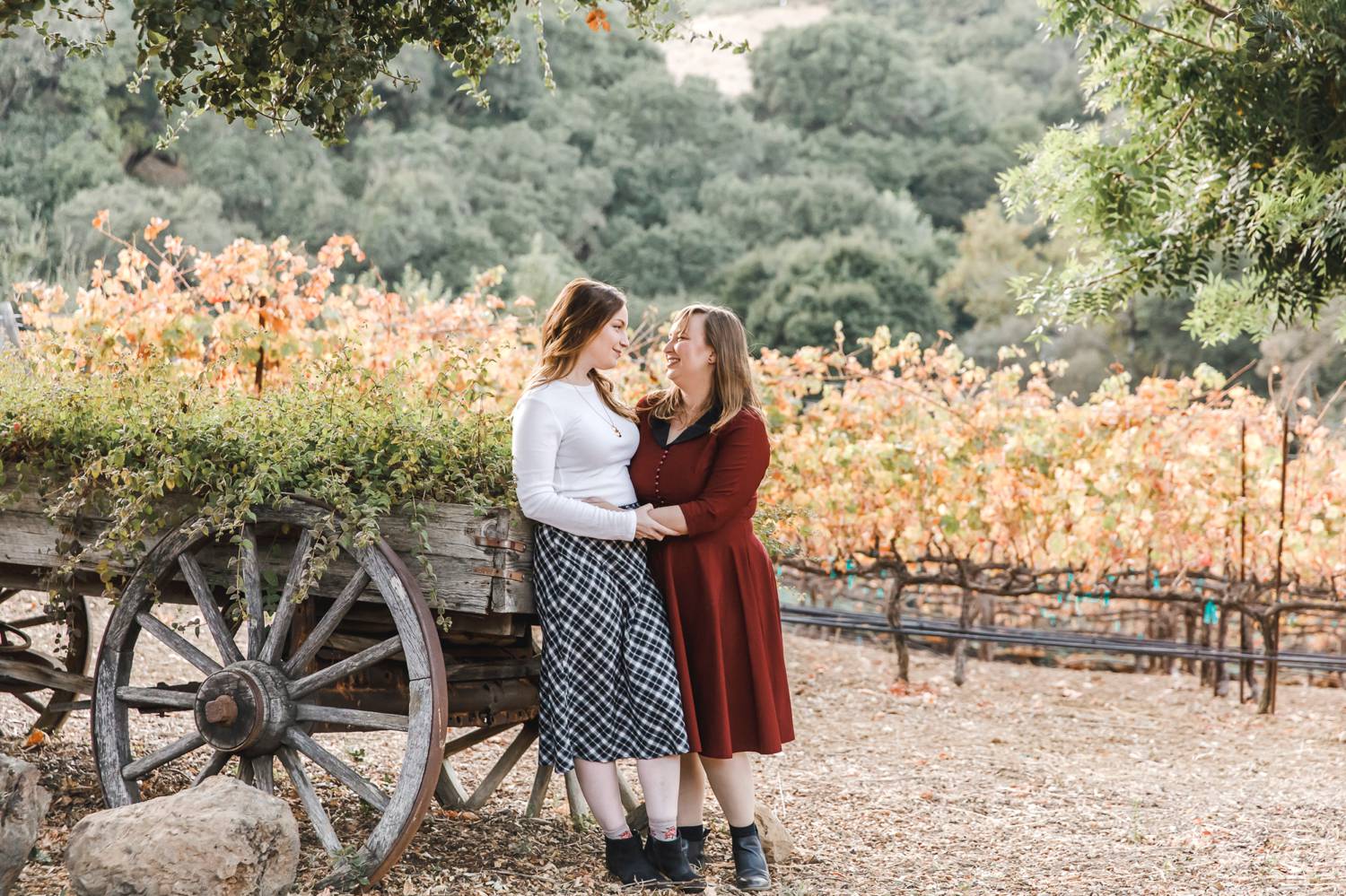 Two women in love stand close together, looking at each other as they lean against an old wagon full of herbs.