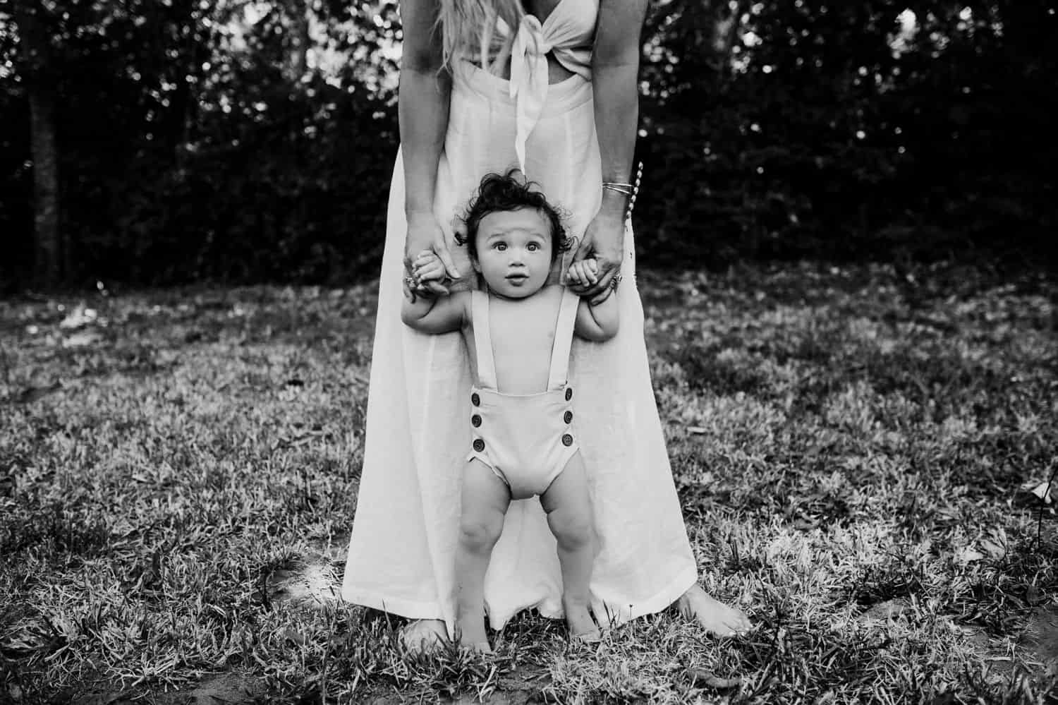 Black and white photo of a toddler standing in front of his mother. He's wearing overalls with no shirt, and only the mother's arms and legs are visible.
