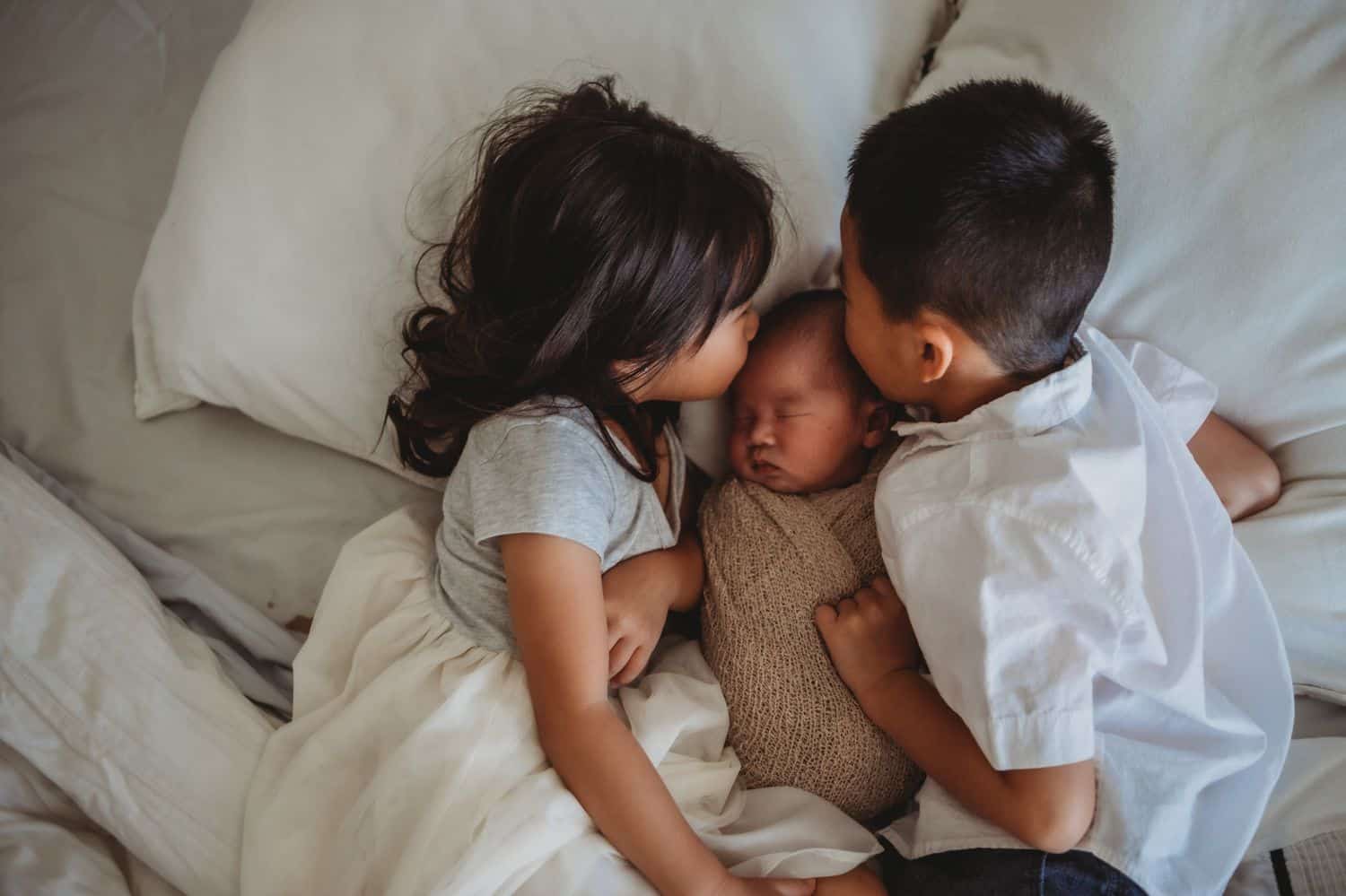 Three children lie together in a bed with ivory linens. The toddler brother and sister kiss their infant sibling on the head.