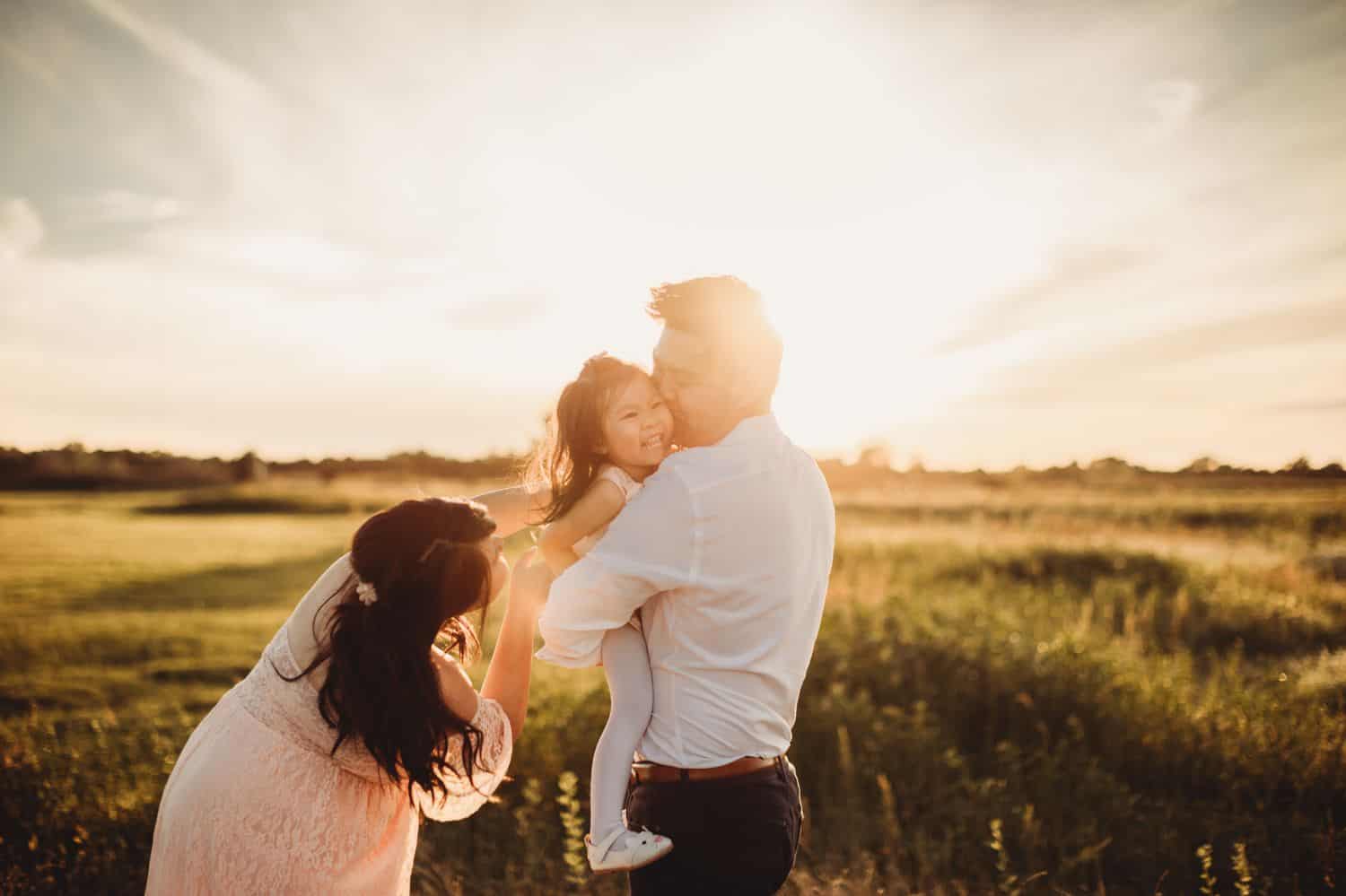 Two parents are surrounded by sun flare as they play with their toddler daughter in a field.