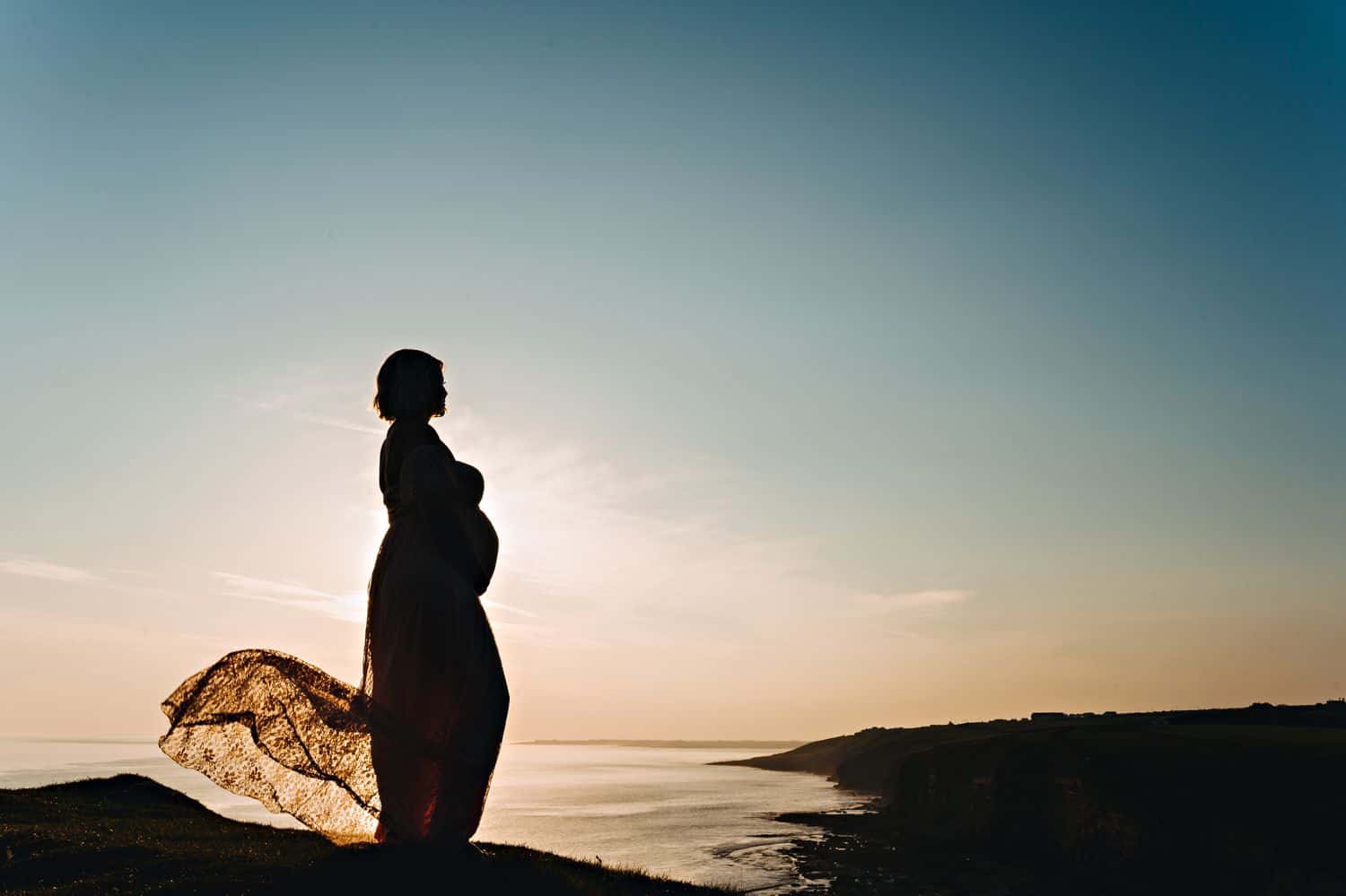 Silhouette of a pregnant woman in a flowing lace dress at the beach at sunset.