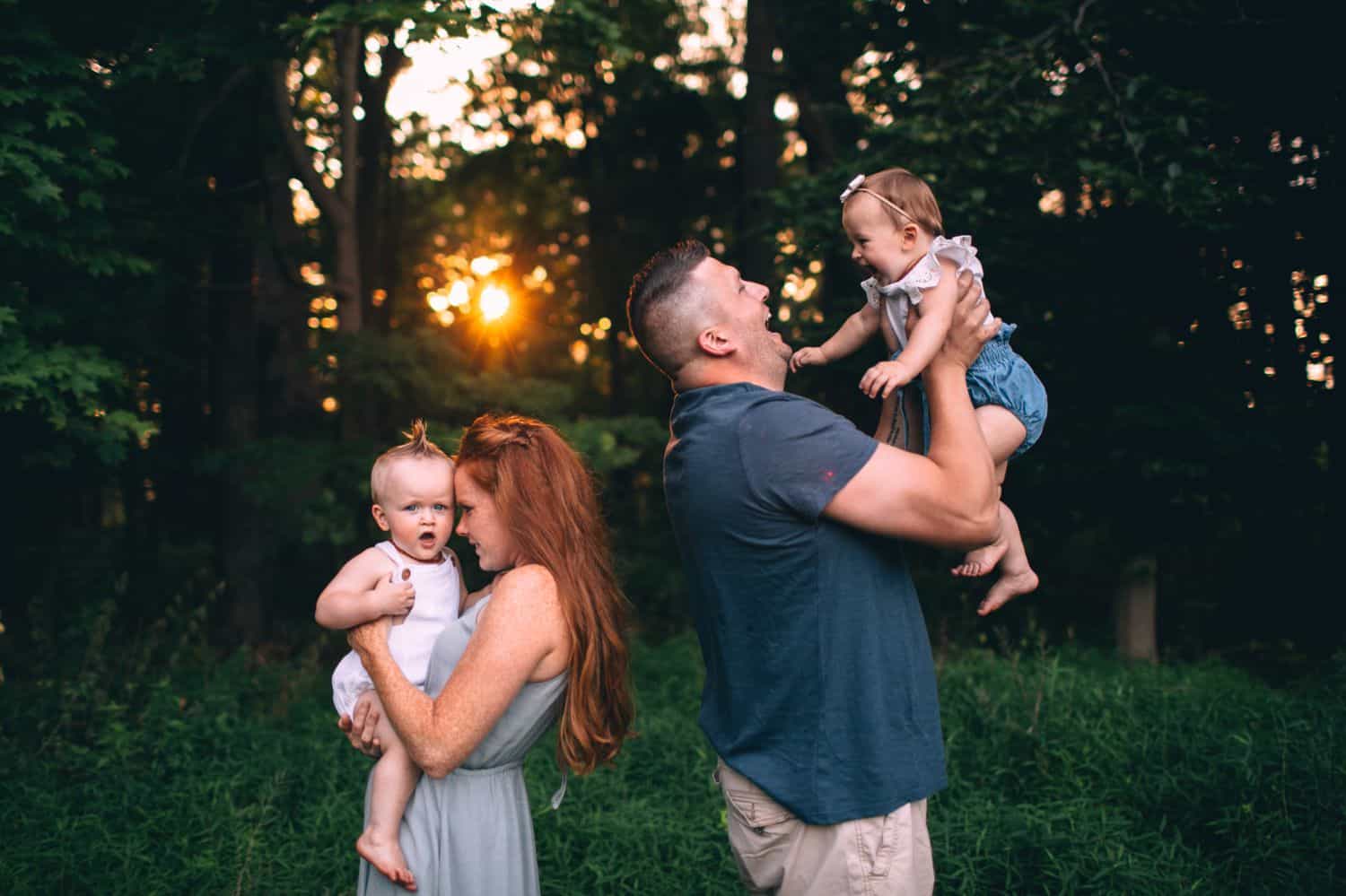 Two parents stand back-to-back at sunset, each holding one of their toddler children against a forest backdrop.