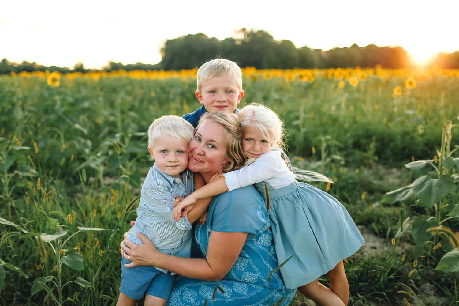 A mother kneels in a corn field at sunset. She is surrounded by her three young children.