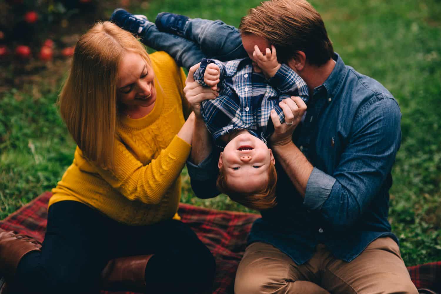 Two parents dressed in fall colors sit on a plaid picnic blanket. They are holding their toddler son upside down as he giggles.