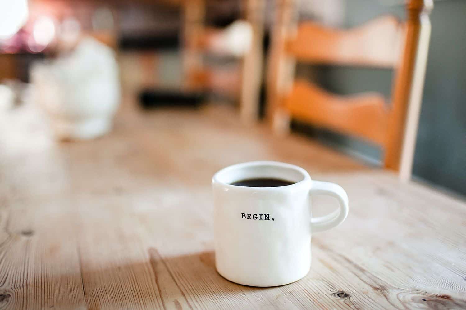 A white mug of coffee with the word "Begin" engraved on the side sits on a plain wooden table.