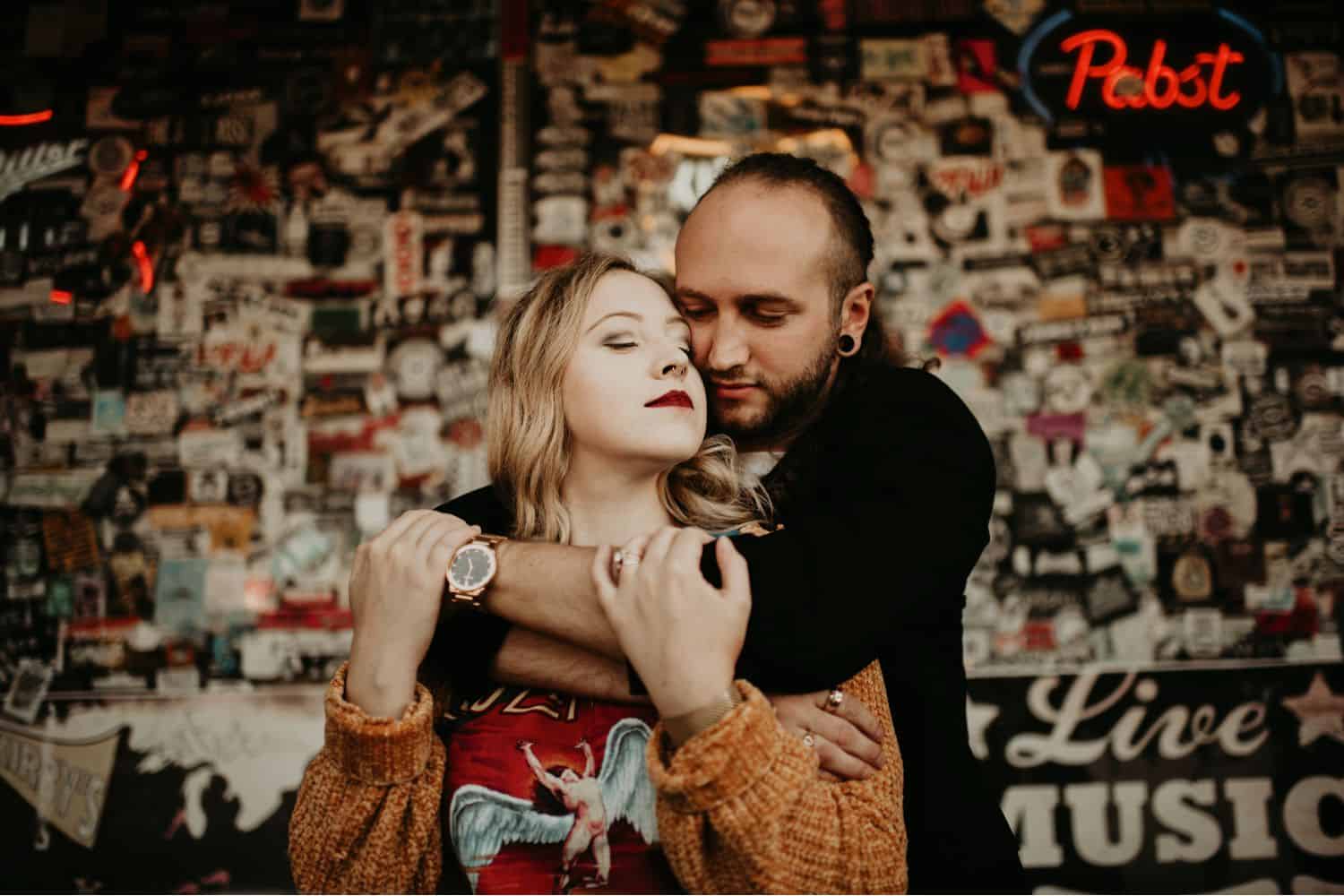 A hipster couple embraces in a bar in front of a wall full of band stickers. Learn how to take low light photos like this one from Shelby Laine Photography