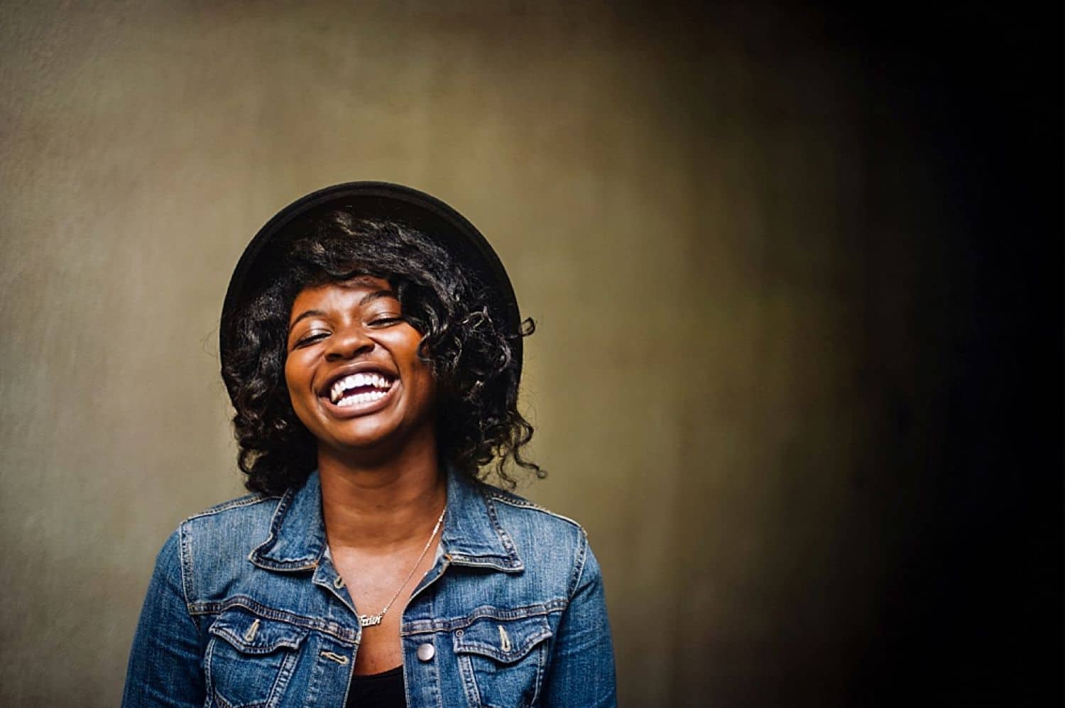 A young woman wearing a hat and a denim jacket smiles brightly with her face tilted toward the sky. By Kate T. Parker for Strong is the New Pretty