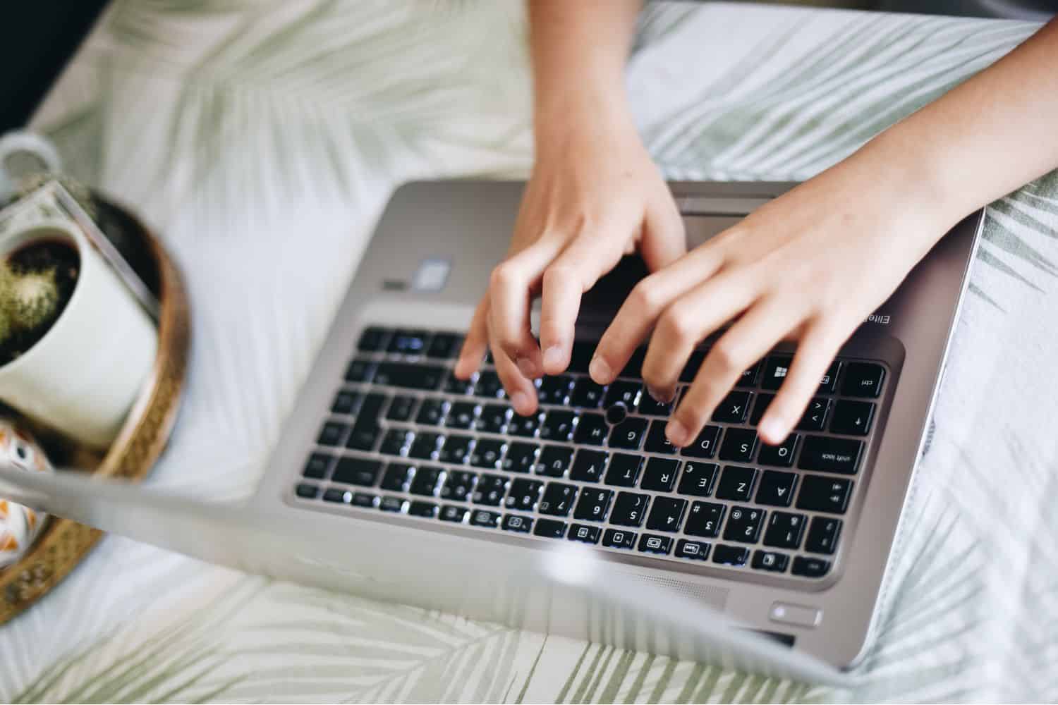 Hands type on a laptop that rests on a fern-printed comforter. Photo by Gaelle Marcel.