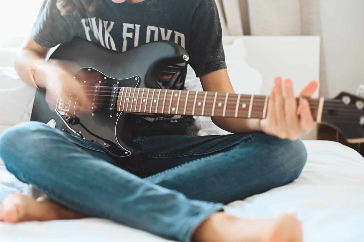 A person sits cross-legged on a white bed playing an electric guitar.