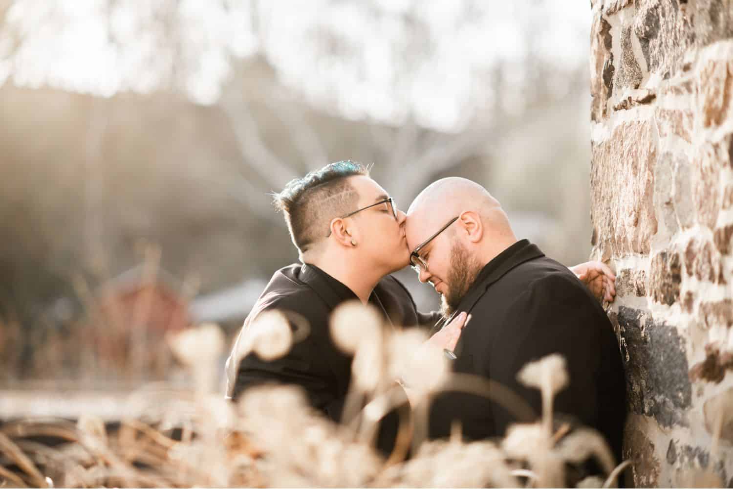 A groom kisses his husband's forehead as they stand outside against a stone wall