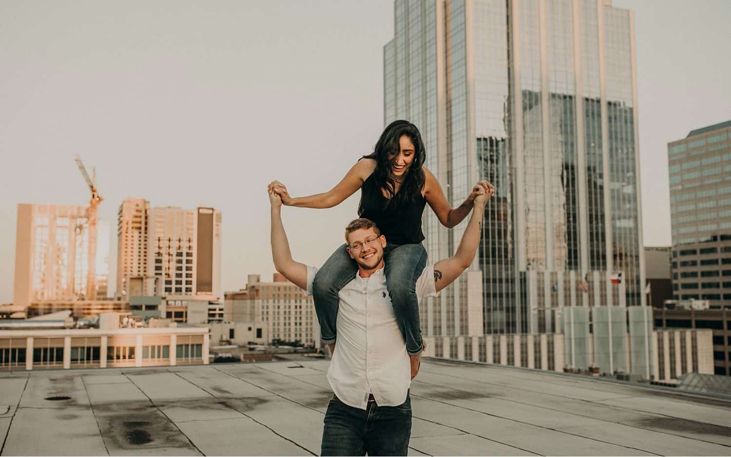 A woman sits on her boyfriend's shoulders and holds his outstretched hands as he walks across a city rooftop
