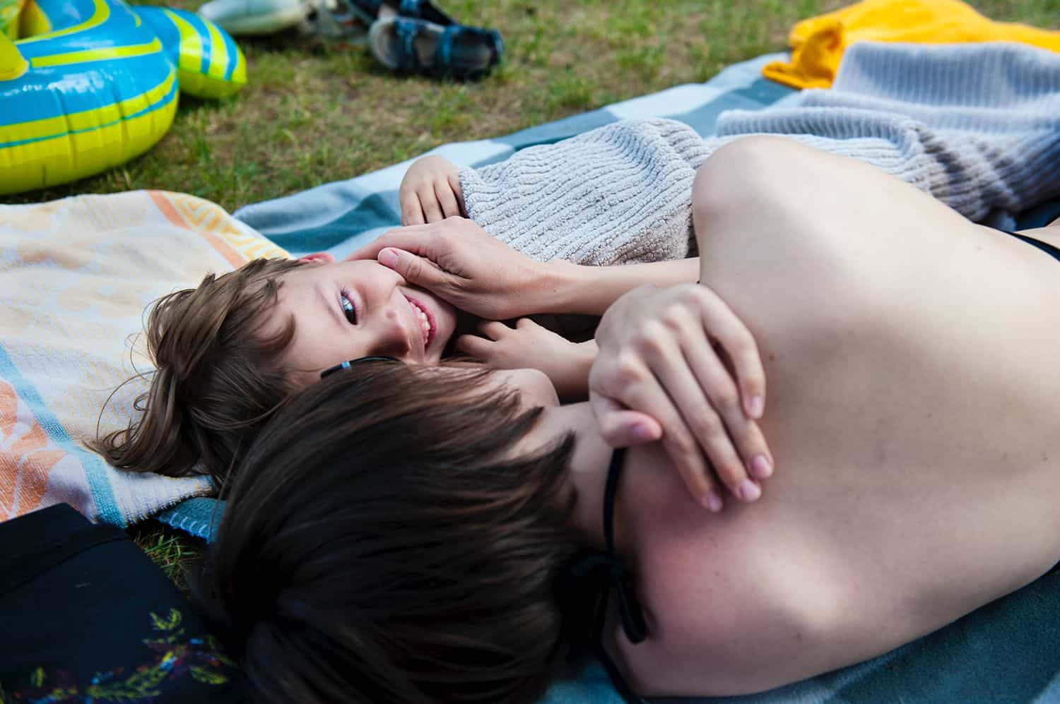 A mom and her child snuggle close while lying in bathing suits on a massive beach blanket