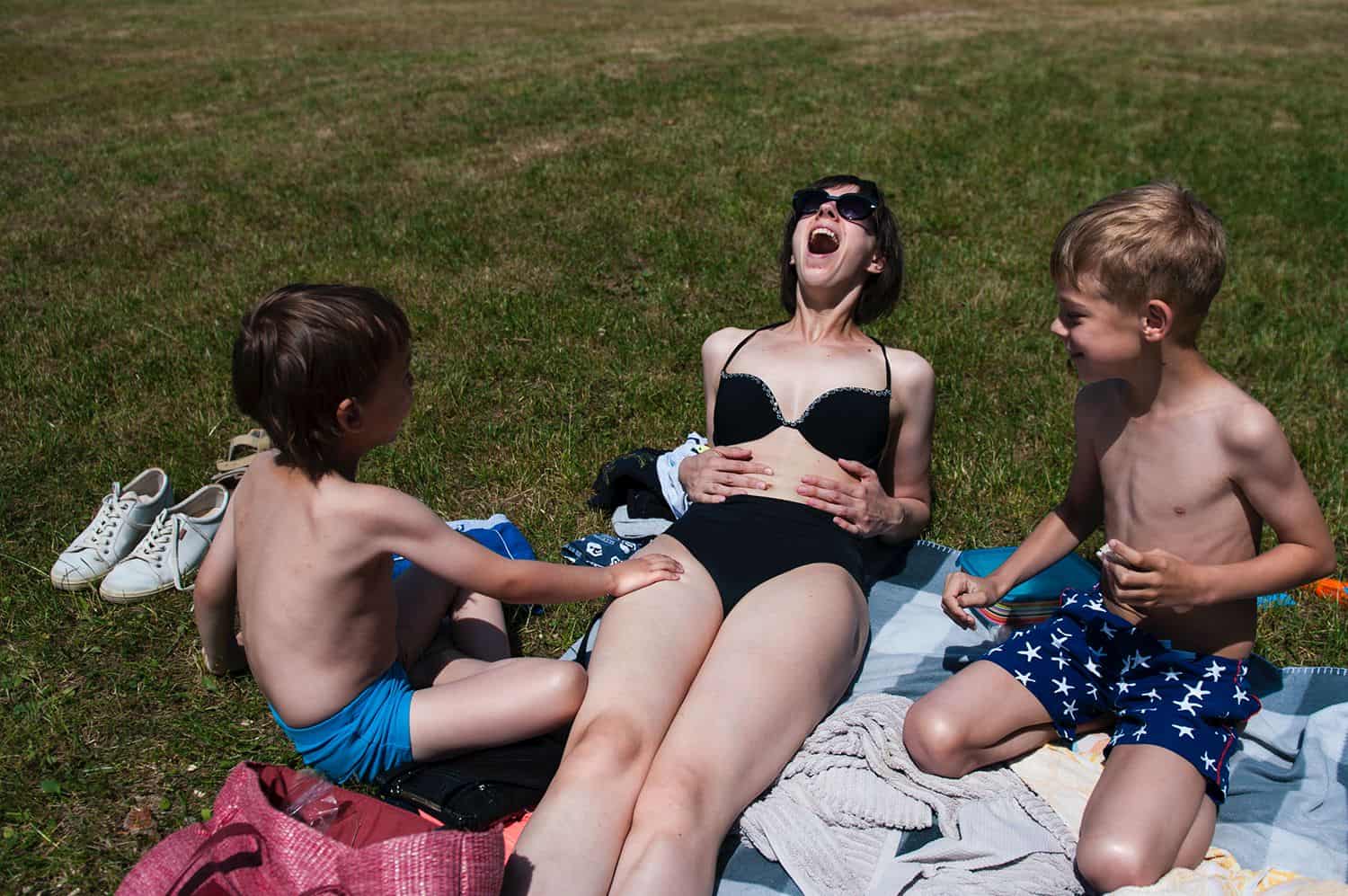 A mom in a black bikini and sunglasses lies on beach blankets with her sons and laughs wildly