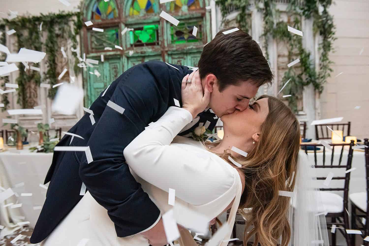 A groom dips a bride for a kiss as confetti rains down on them