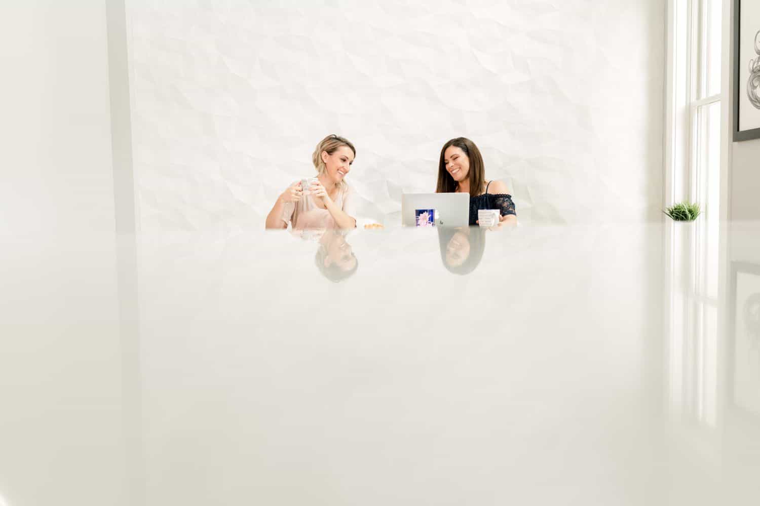 Two business women sit at a large reflective desk in a white room