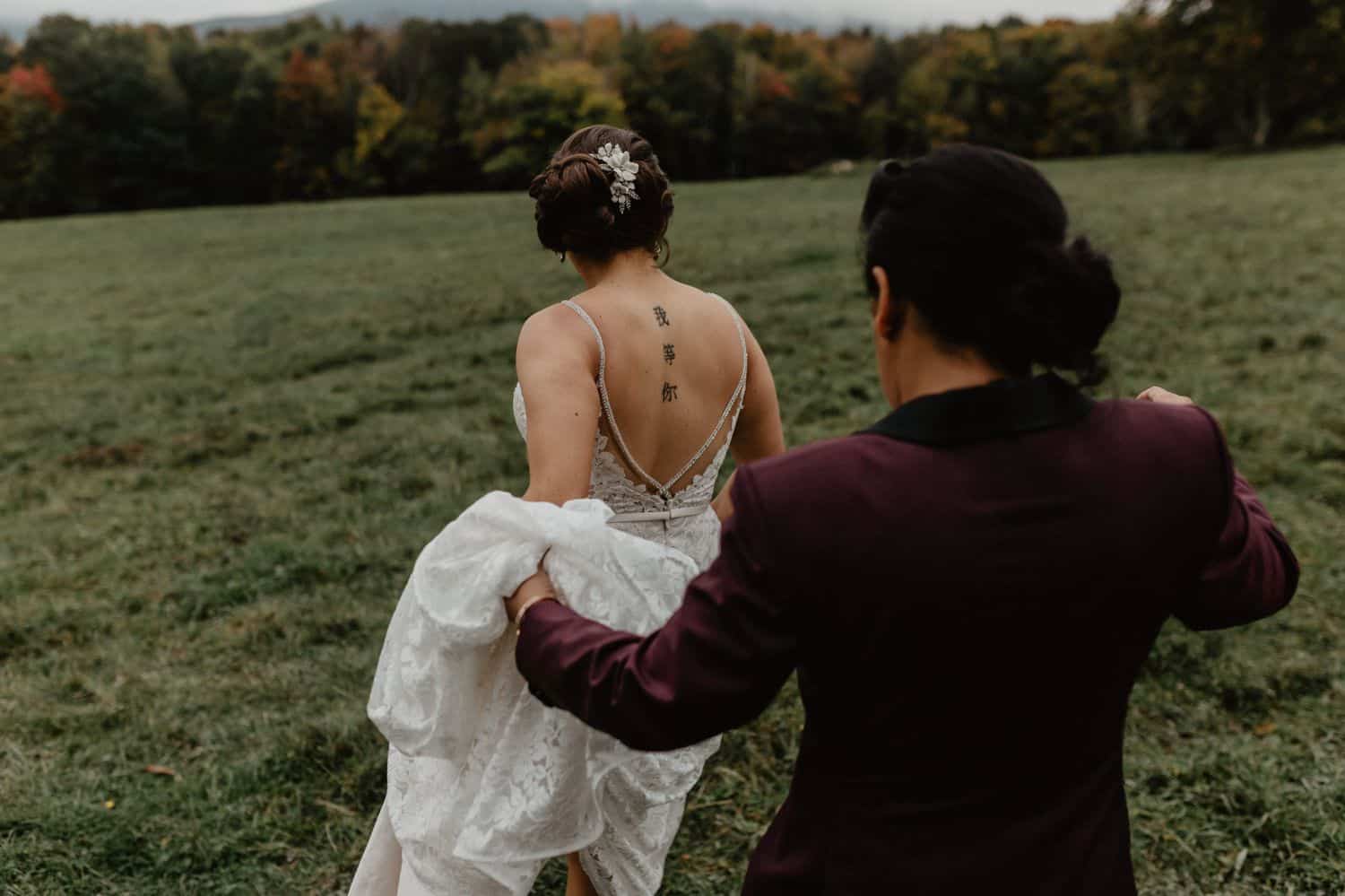 A bride with a back tattoo walks through a field as her wife holds her train.
