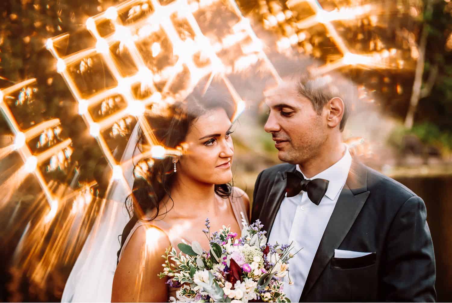 Bride holding bouquet looks at groom with flares of golden light spilling around them.