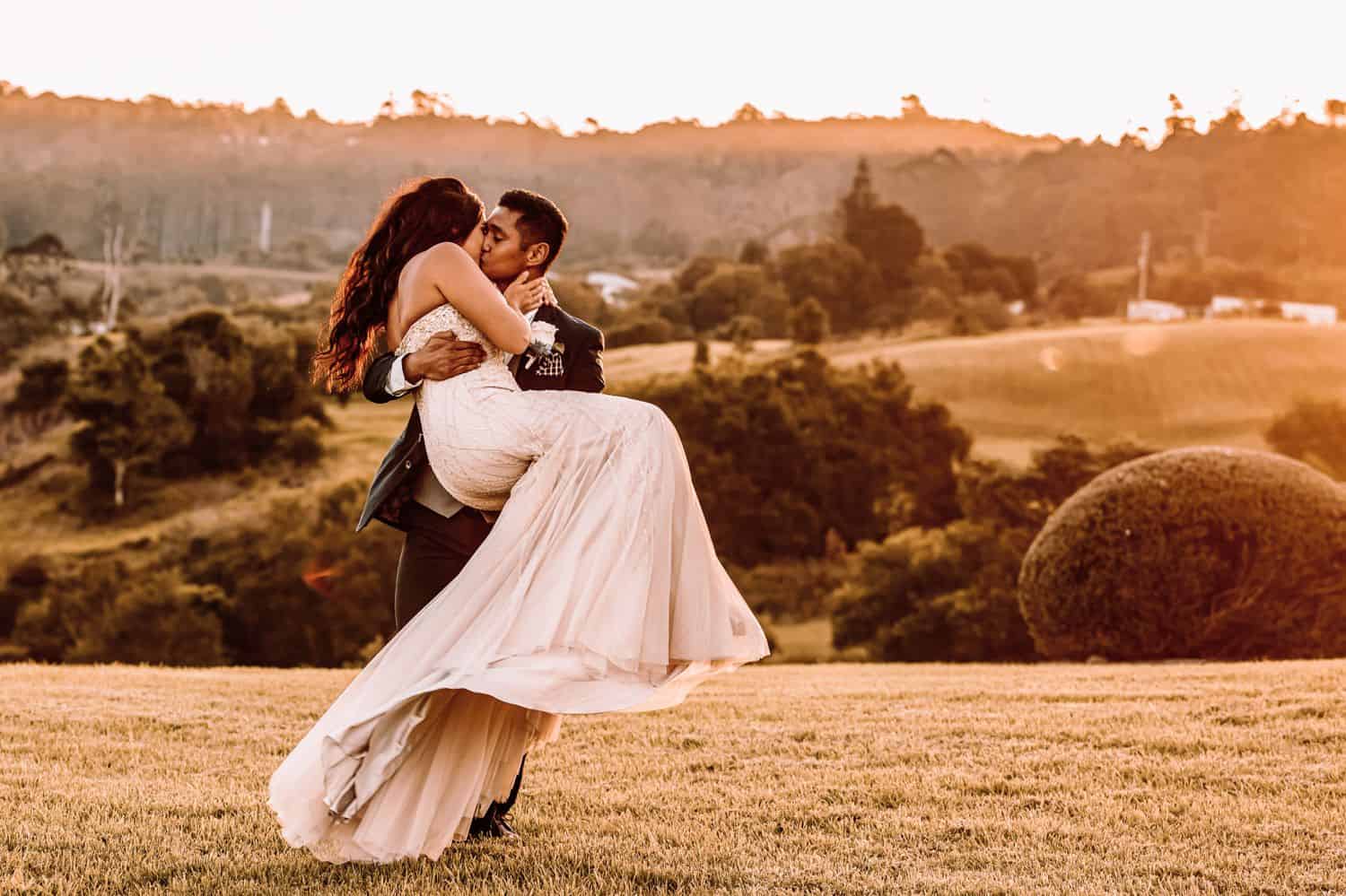 Groom carries bride in a flowing dress in a cradle-hold while they kiss.