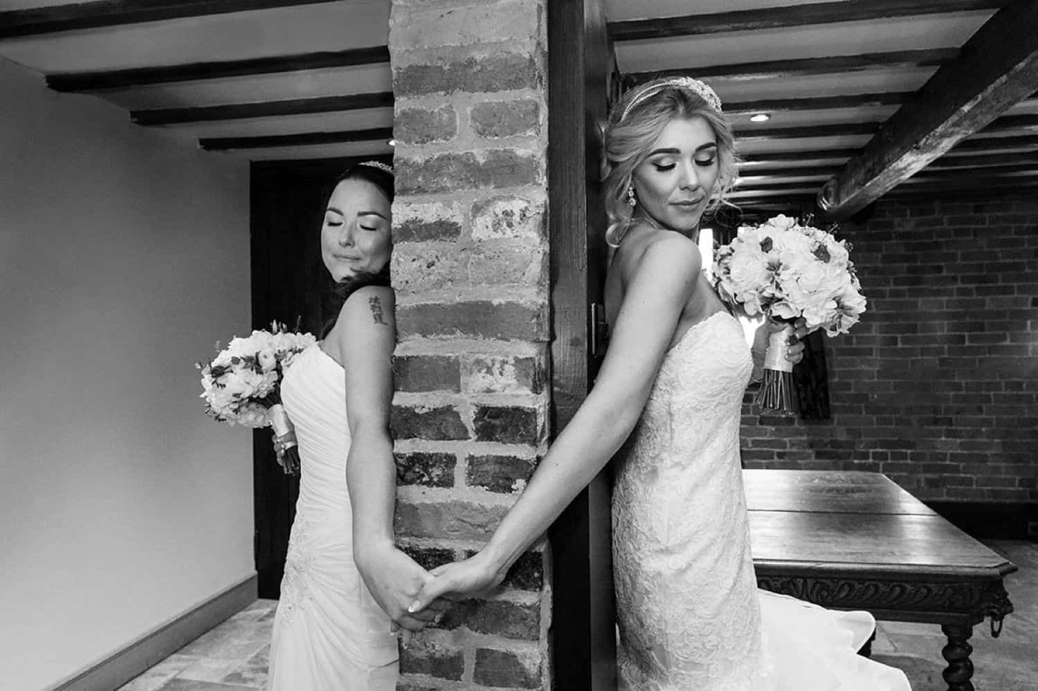 Two brides hold hands on either side of a brick wall before their first look