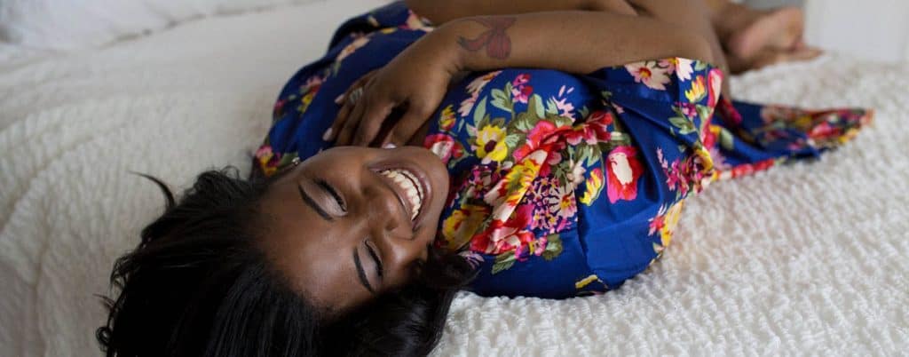 A smiling Black woman lies on a white bed in a blue floral robe for her boudoir session with Kinzie Ferguson of the Empowerment Studio