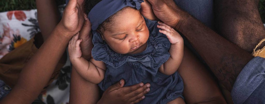 Parallel Summit Creative: A newborn baby girl in a denim dress is cradled by the hands of her family.