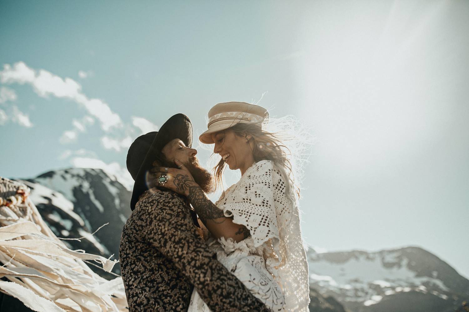 A man in a black hat lifts a woman in a white hat as they smile and hug. Behind them are snow covered mountains.