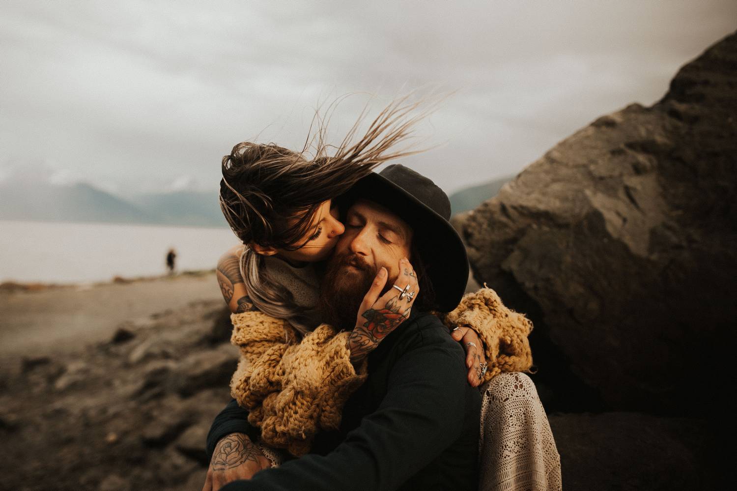 A man in a black hat sits on rocks by the ocean. A woman with her hair blowing in the wind wraps her arms around him and kisses him.