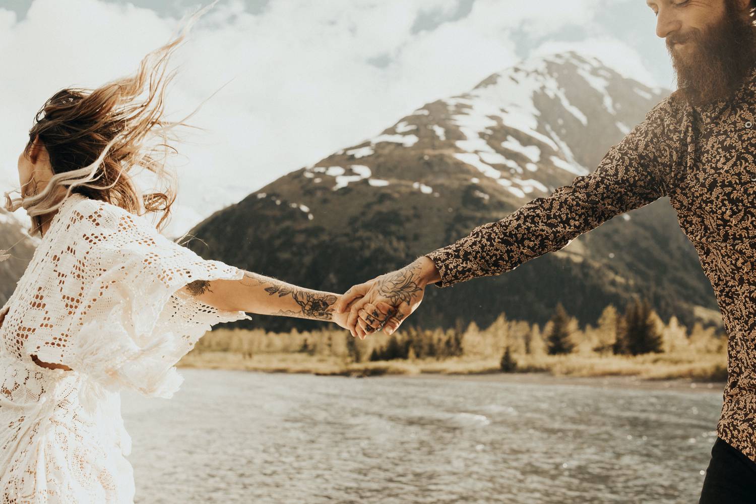 A woman wearing white lace leads a tattooed man toward a mountain lake