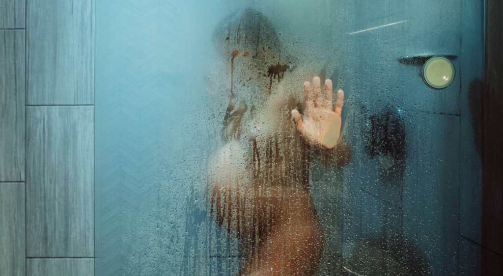 A pregnant person is seen through the fog of a steamy shower door, her hand flat against the glass as she labors.