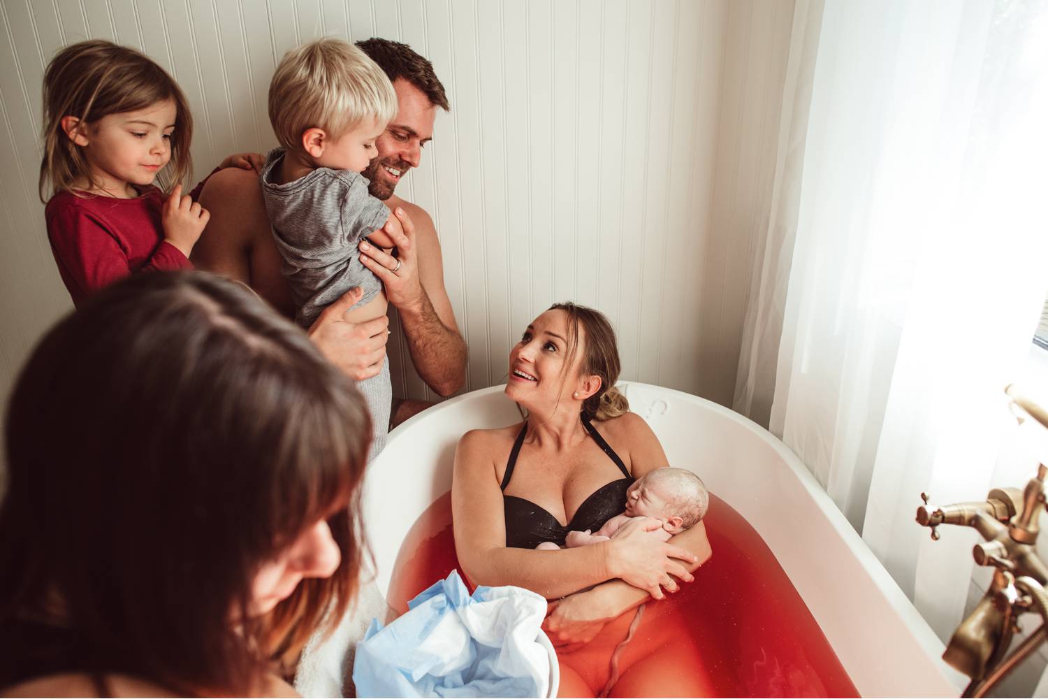 A woman sits in a tub full of red-tinged water holding the new baby she just birthed as her partner and children look on