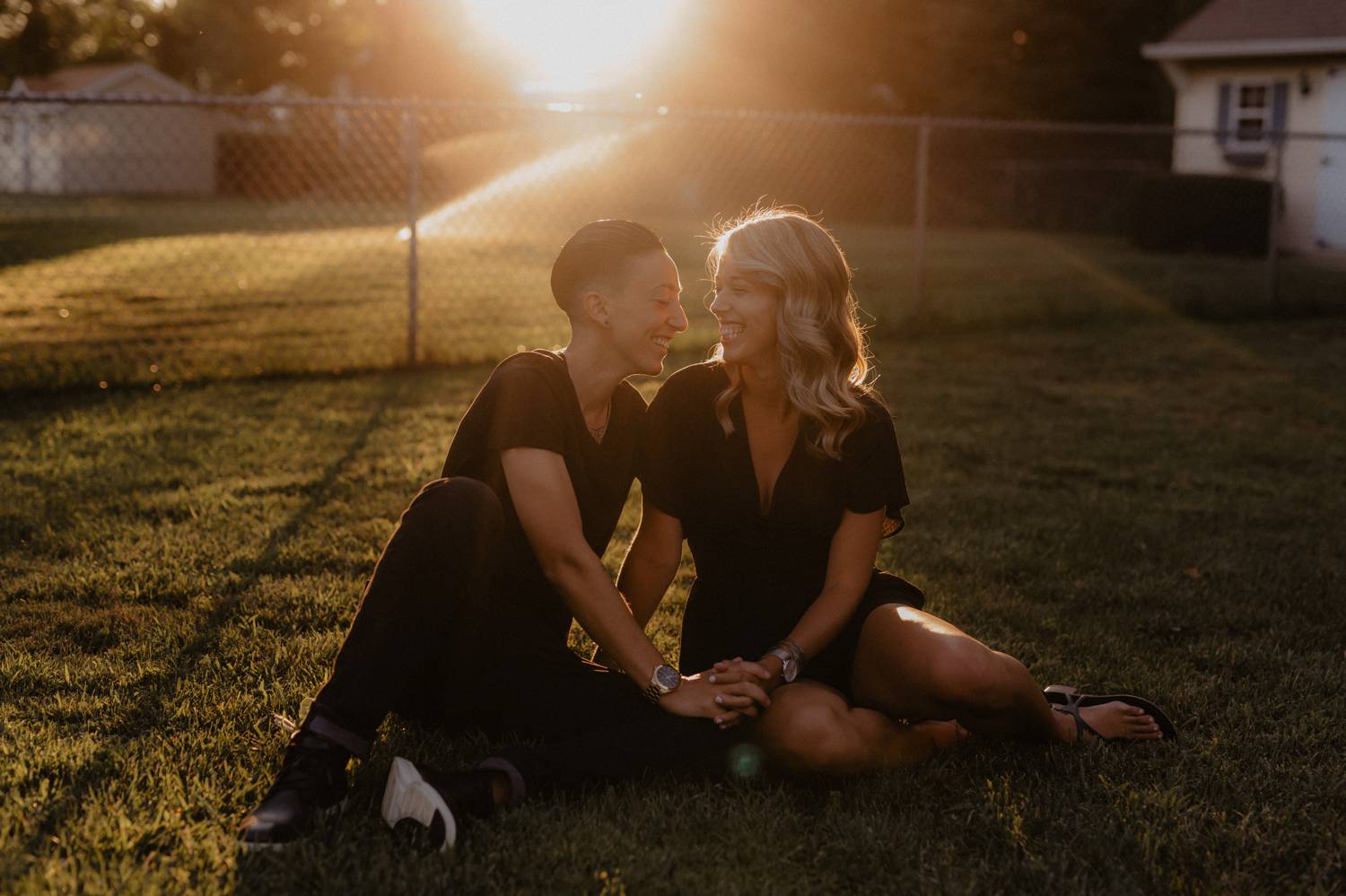 A queer couples sits in the grass as the sun sets, their foreheads nearly touching