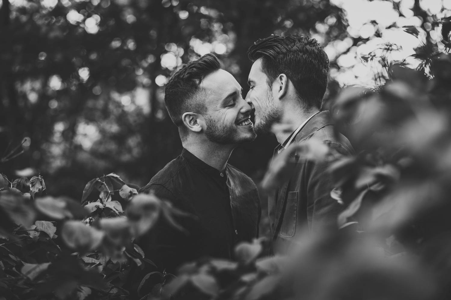 Two engaged men move in two kiss while being photographed through beautiful spring foliage