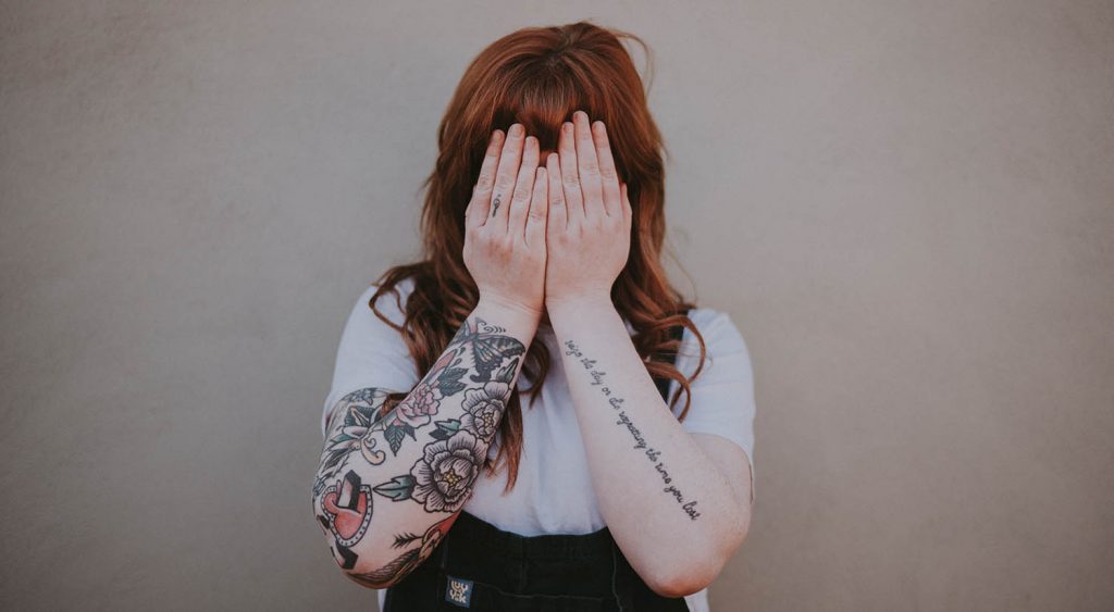 Woman with red hair and tattoos hides her face with her hands. By Annie Spratt photographer