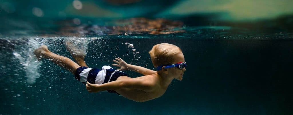 A child wearing striped swim trunks and goggles is swimming underwater with his arms stretched behind him