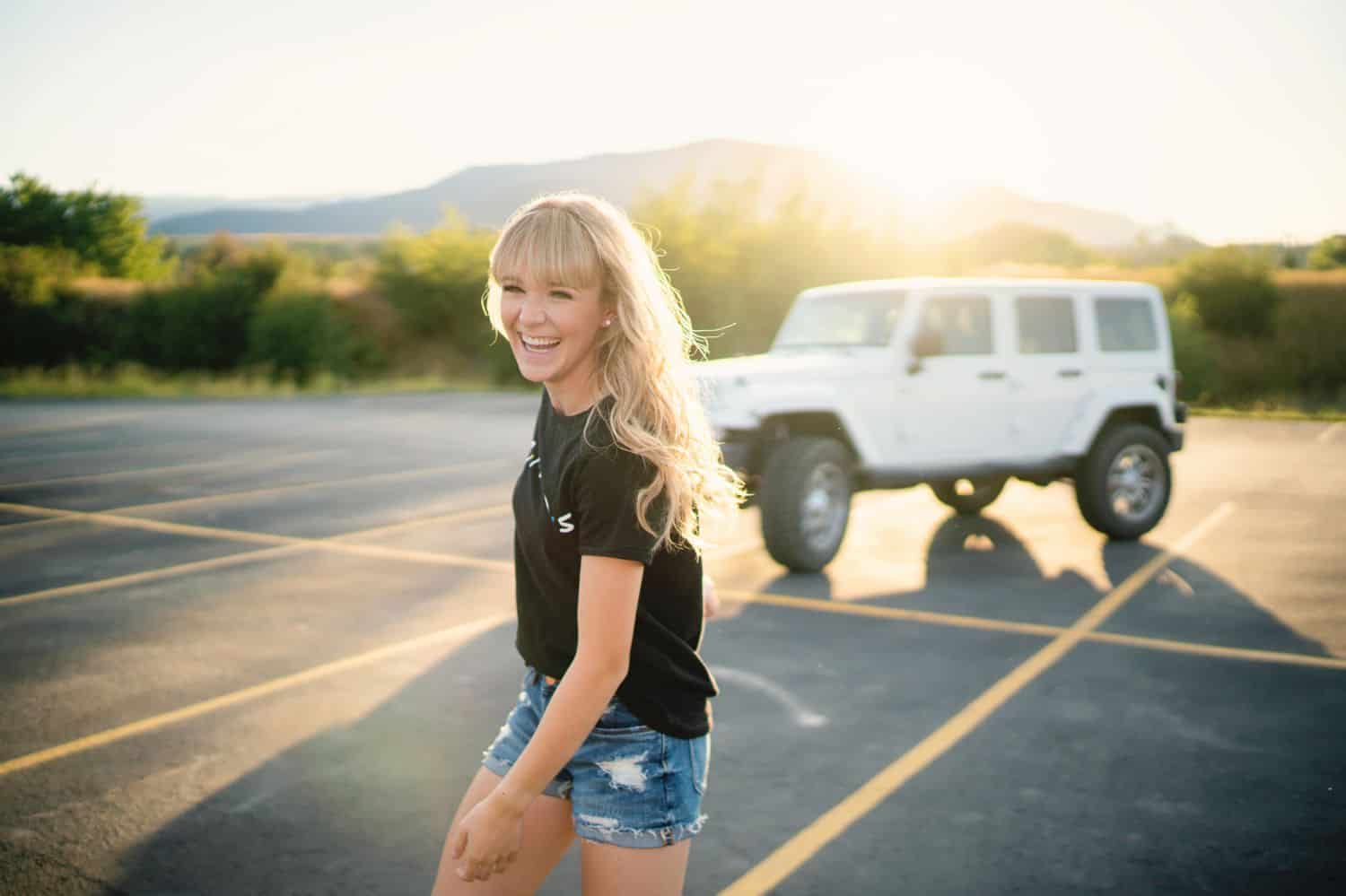 A senior girls smiles as she walks away from her white Jeep parked in the sunset