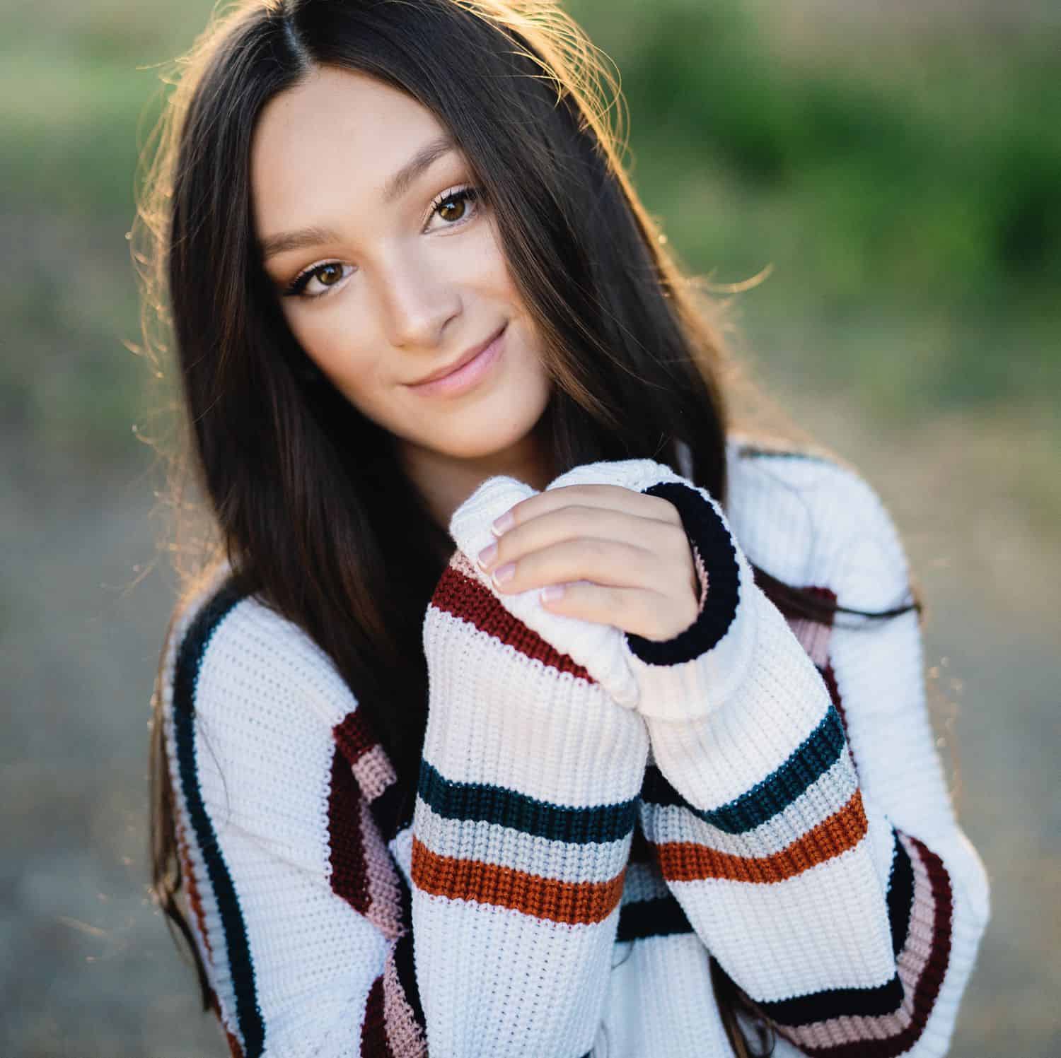 A senior girl in a striped sweater stands backlit in a field with her hands clasped by her face